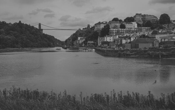 A view of the River Avon at high tide, with Hotwells and the Clifton Suspension Bridge in the background as recreational boats approach Entrance Lock on a grey, flat day.